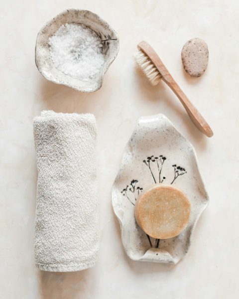 Bath salt, a wooden brush, a rolled towel and a clay bar on a speckled ceramic dish