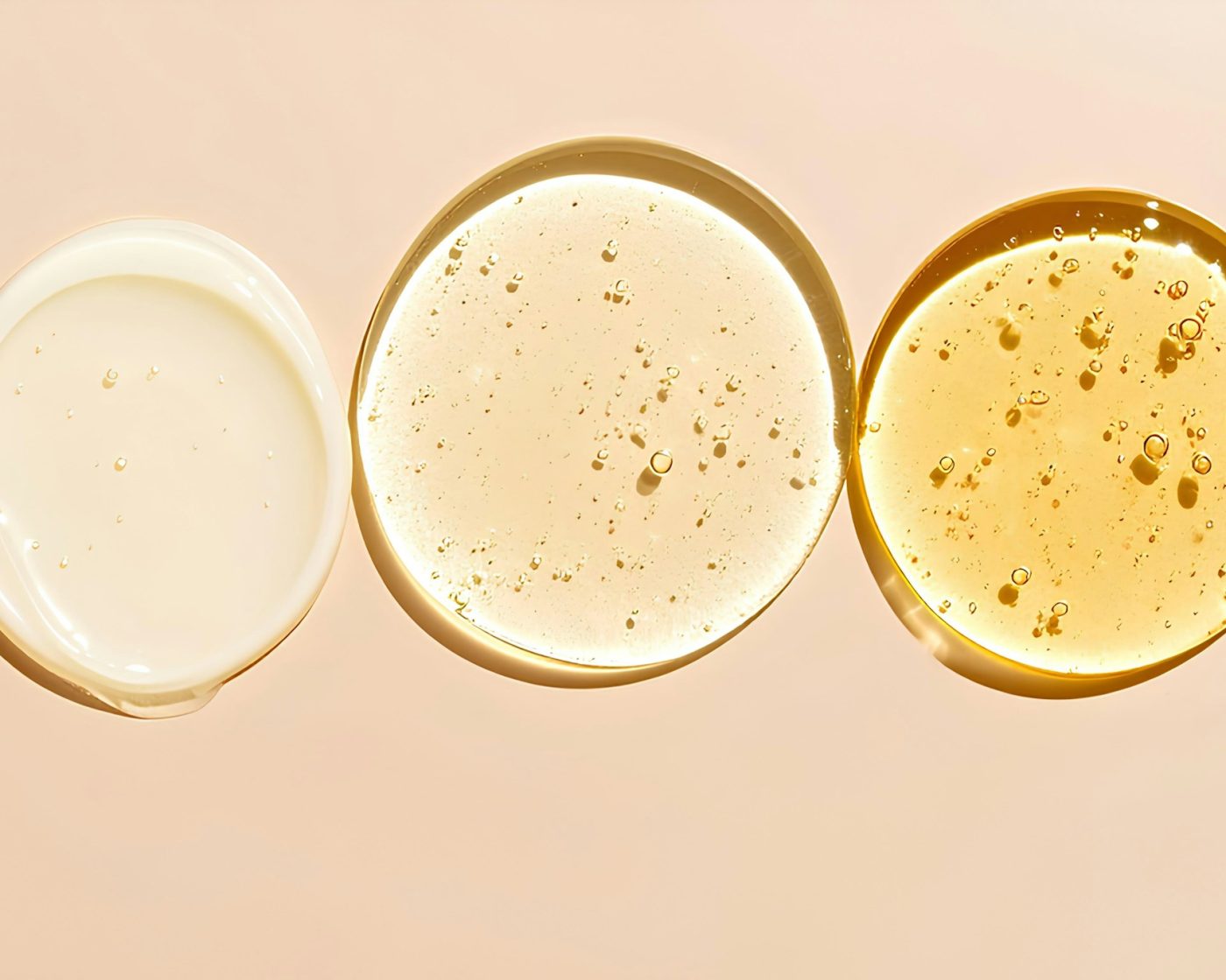 Swatches of a white cream, a clear serum and a golden oil on a soft peach background