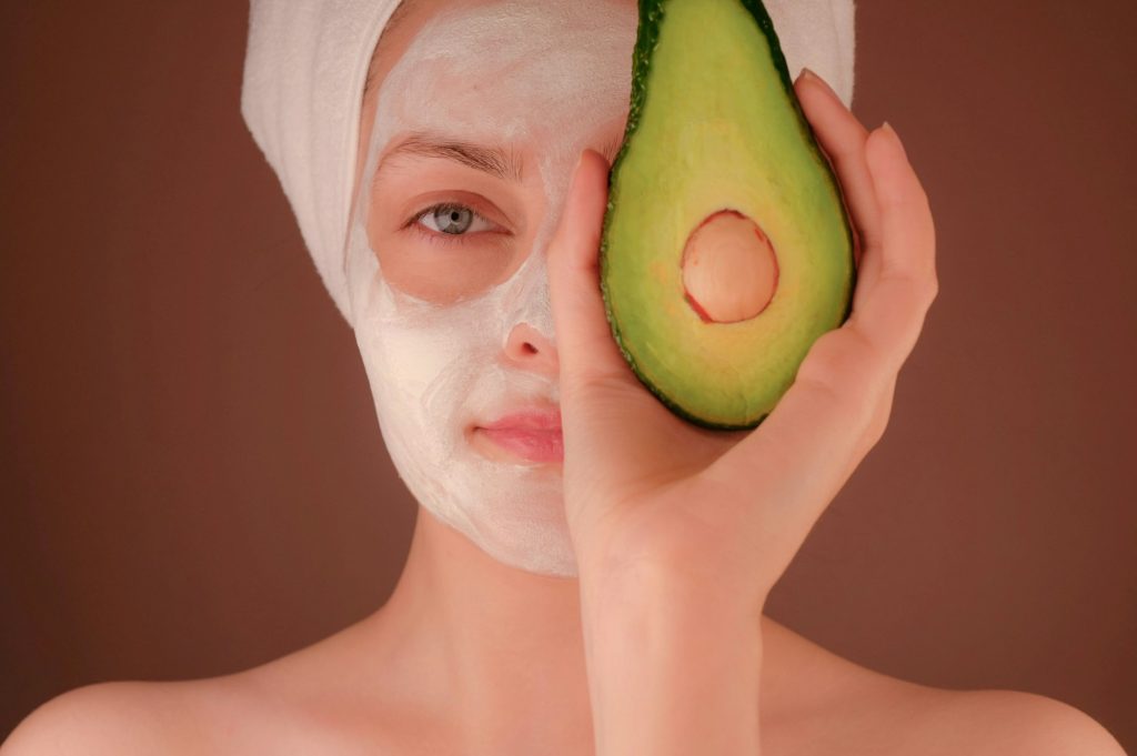 A woman wearing a pale clay face mask, holding half an avocado