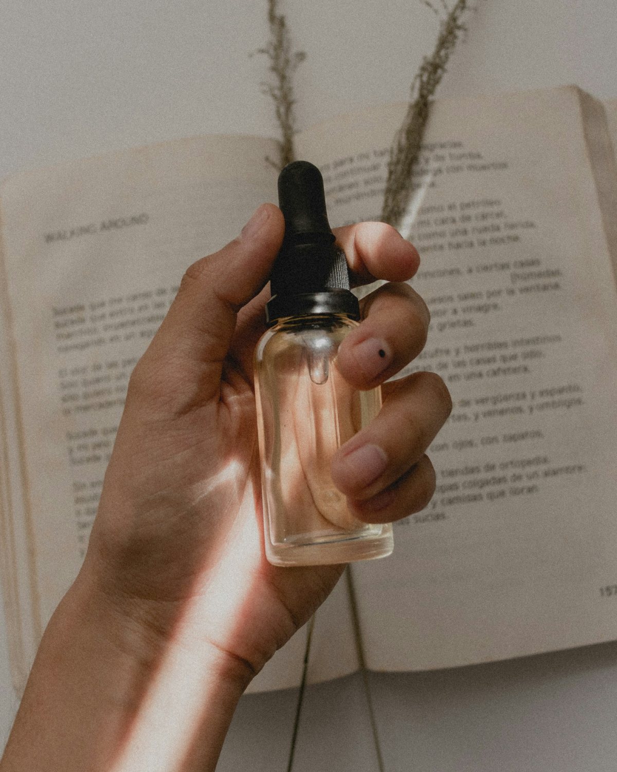 Hand holding a clear dropper bottle over an open book and dried grasses