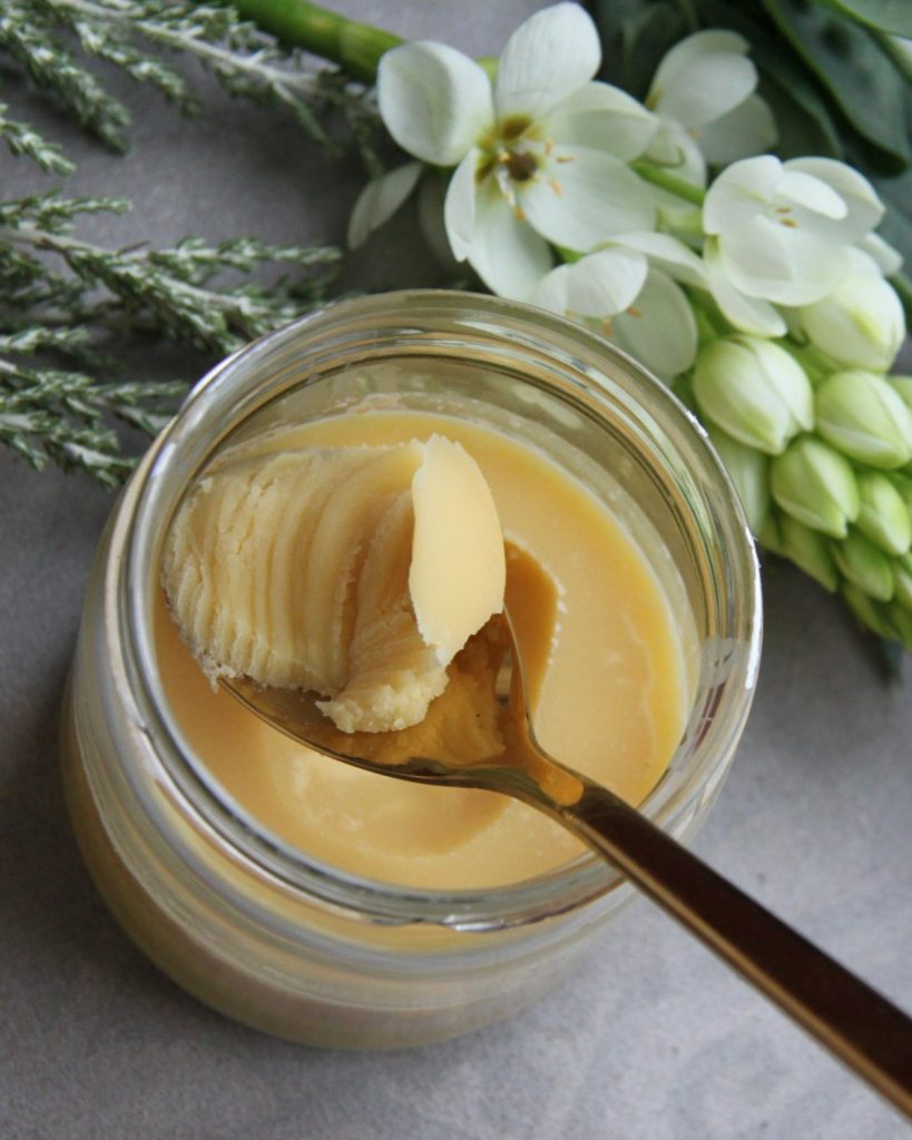 Glass jar of whipped body butter with a gold spoon beside fresh flowers