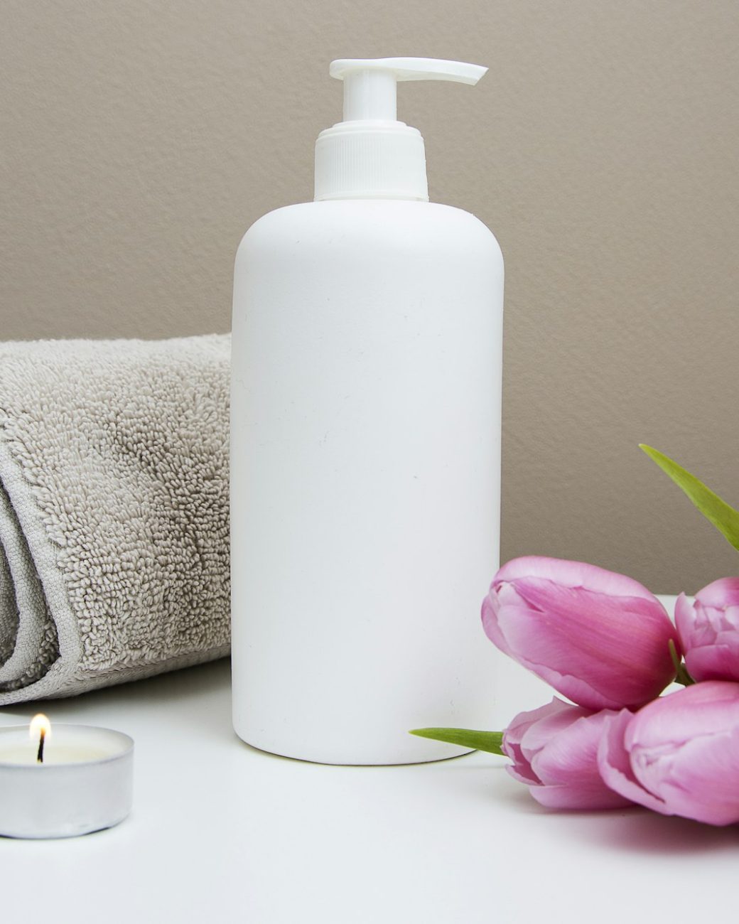 White pump bottle beside a rolled grey towel, pink tulips and a tea light